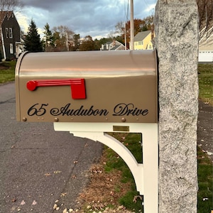 May include: A brown mailbox with a red flag and the address "65 Audubon Drive" in black lettering. The mailbox is mounted on a white bracket attached to a gray granite post.