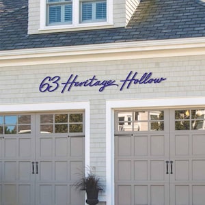 Könnte beinhalten: Ein dunkelblaues Metall-Wandschild mit dem kursiven Schriftzug 63 Heritage Hollow, montiert an der hellgrauen, geschindelten Außenwand eines Hauses über zwei grauen Garagentoren mit rechteckigen Fensterscheiben und schwarzen Griffen.
