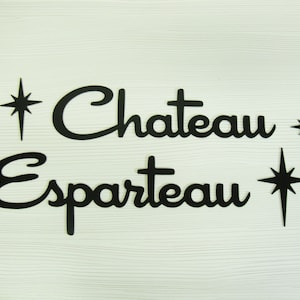 Könnte beinhalten: Schwarze ausgeschnittene Buchstaben, die "Chateau Esparteau" buchstabieren, mit drei schwarzen Sternformen auf einem weißen Holzgrund.
