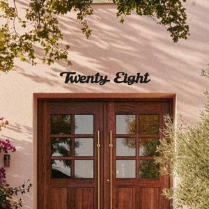 May include: A wooden double door with a grid pattern, featuring gold handles, set against a light pink wall. The words "Twenty Eight" are written in a cursive font above the door. Green foliage frames the scene.