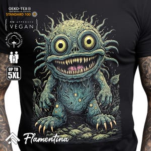 May include: Black t-shirt featuring a green and blue monster graphic. The monster has a wide grin with sharp teeth and large eyes. The text "Flamentina" is printed below the monster.
