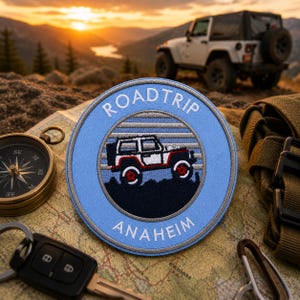 May include: A round, light blue patch with the words "ROADTRIP" and "ANAHEIM" embroidered in white. The patch features a graphic of a white and red jeep. A compass, keys, and a map are also visible.