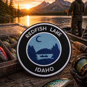 Op de afbeelding: Een ronde Redfish Lake Idaho patch met een blauw en wit ontwerp van een meer en bergen. De patch ligt op een houten oppervlak met kunstaas en visgerei. Op de achtergrond vist een persoon.
