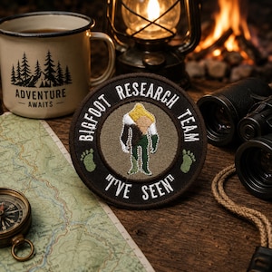 Può includere: Una toppa ricamata circolare raffigurante una figura di Bigfoot marrone, bianca e gialla su sfondo marrone chiaro. Il bordo nero contiene il testo bianco BIGFOOT RESEARCH TEAM e I'VE SEEN, accentuato da due piccole impronte verdi sui lati.