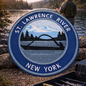 May include: A circular patch with the text "ST. LAWRENCE RIVER NEW YORK" in white lettering. The patch features a blue border and a central design of a bridge, mountains, and trees. A person is fishing in the background.