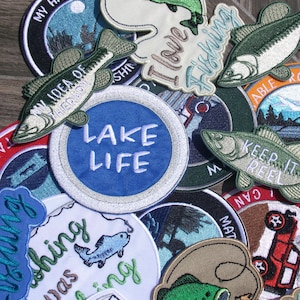 May include: A collection of embroidered patches with fishing and lake life themes. Designs include fish, the text "Lake Life", and phrases like "I love fishing" and "Keep it Reel". Patches are in various colors, including blue, green, and white.