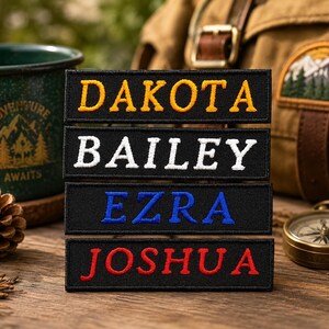 May include: Four rectangular black embroidered patches stacked vertically. Each patch features a name in capital letters: DAKOTA in yellow, BAILEY in white, EZRA in blue, and JOSHUA in red. The patches have a stitched border and a fabric texture.
