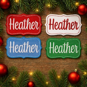 Peut inclure: Quatre écussons brodés avec le nom "Heather" en écriture blanche. Les écussons sont rouges, blancs, bleus et verts, chacun avec une bordure décorative. Le fond comprend des ornements de Noël et des branches de pin.