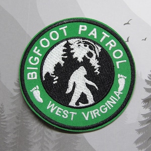 Könnte beinhalten: Ein gestickter Aufnäher in Grün und Weiß mit dem Text "Bigfoot Patrol West Virginia" und einer Silhouette von Bigfoot, der vor einem Wald geht.