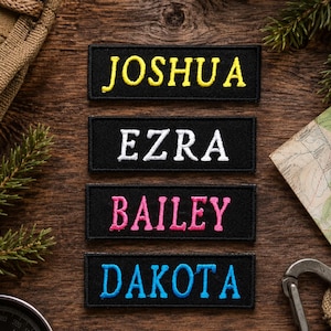May include: Four black rectangular patches with embroidered names: JOSHUA in yellow, EZRA in white, BAILEY in pink, and DAKOTA in blue. The patches are arranged on a wooden surface, with a compass, map, and carabiner visible.