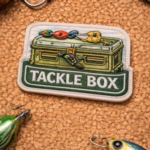May include: An embroidered patch featuring a green tackle box with the words "TACKLE BOX" in white. The patch is surrounded by fishing lures and tools, set against a textured, brown background.