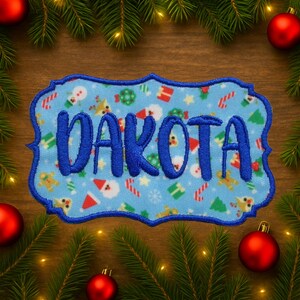 May include: A light blue embroidered patch with the name "DAKOTA" in bold blue letters. The patch has a decorative border and is decorated with Christmas-themed patterns. The patch is surrounded by Christmas decorations.