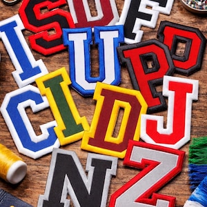 May include: Assorted embroidered varsity letter patches in various colors, including red, blue, and yellow, with white borders. The letters are arranged on a wooden surface, alongside sewing supplies like scissors and thread.