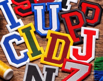 May include: Assorted embroidered varsity letter patches in various colors, including red, blue, and yellow, with white borders. The letters are arranged on a wooden surface, alongside sewing supplies like scissors and thread.