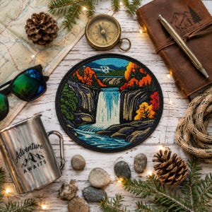 May include: A circular embroidered patch featuring a landscape with a waterfall, dark cliffs, and autumn-coloured trees in shades of orange and yellow. The patch is surrounded by a pinecone, a compass, a map, a metal mug, and a leather journal.