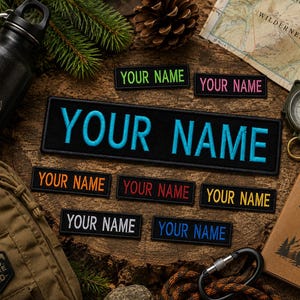 May include: A collection of rectangular black fabric patches featuring the text YOUR NAME embroidered in various colors including bright blue, lime green, pink, orange, red, yellow, white, and dark blue, arranged on a rustic wooden surface.