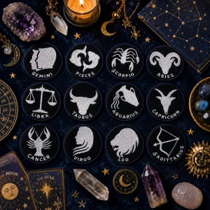 May include: A collection of twelve black circular patches, each embroidered with a white zodiac symbol and name. The image includes tarot cards, crystals, a lit candle, and celestial-themed jewelry, creating an astrological theme.