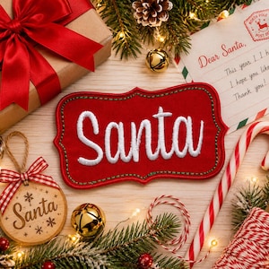 Puede incluir: Un parche de tela roja con borde festoneado y la palabra Santa bordada en hilo blanco. El parche presenta costuras verdes a lo largo del borde y está rodeado por una caja de regalo con un lazo rojo, una carta a Santa y un adorno de madera.