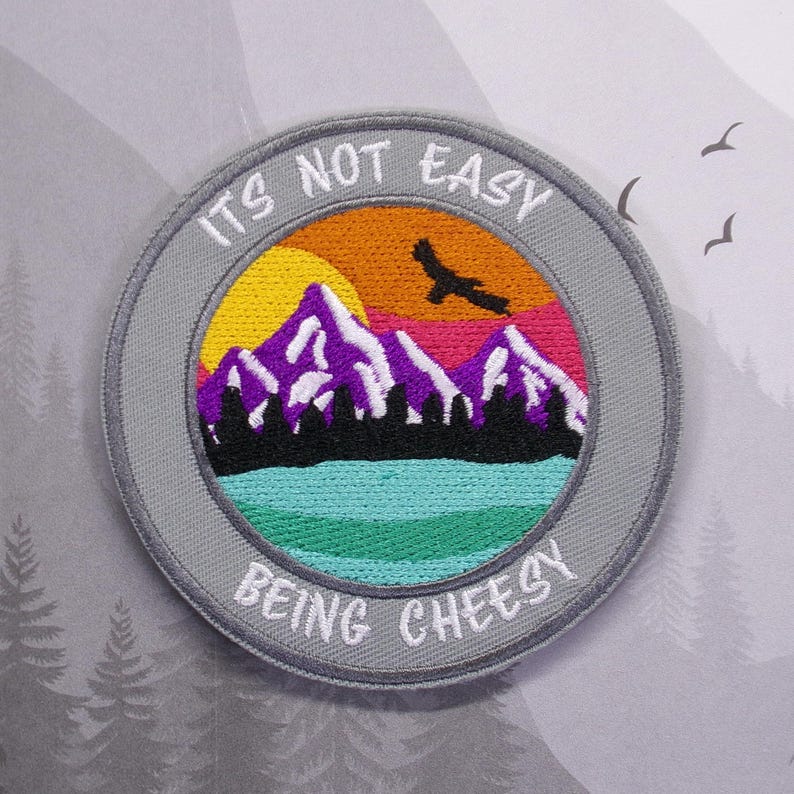 May include: A gray embroidered patch with a mountain scene and a sunset. The text reads "It's not easy being cheesy".
