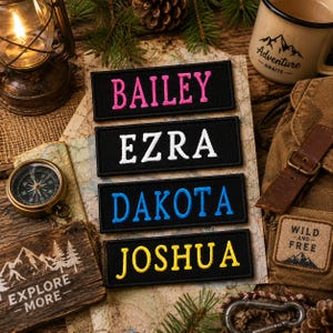 May include: Four black embroidered name patches with the names BAILEY, EZRA, DAKOTA, and JOSHUA in pink, white, blue, and yellow thread. The patches are displayed on a map with a compass, lantern, and a mug that says "Adventure Awaits."
