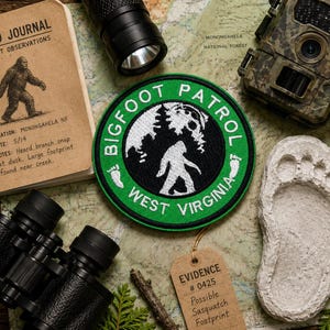 Può includere: Una toppa ricamata circolare con bordo verde e testo BIGFOOT PATROL WEST VIRGINIA in bianco, che circonda un centro nero con la sagoma bianca di una creatura, alberi di pino e due piccole impronte bianche sui lati.