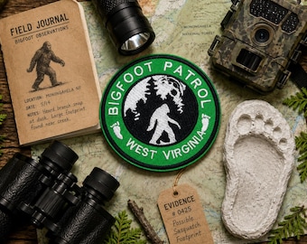 May include: A circular embroidered patch featuring a green border with the text BIGFOOT PATROL WEST VIRGINIA in white, surrounding a black center with a white silhouette of a creature, pine trees, and two small white footprints on the sides.