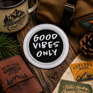Puede incluir: Un parche bordado en blanco y negro con las palabras "GOOD VIBES ONLY". La imagen también presenta una taza con el texto "Adventure Awaits", un diario de cuero y tarjetas temáticas de viaje.