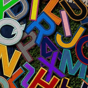 May include: A vibrant assortment of embroidered letter patches in various colors, including blue, yellow, orange, and purple. The patches are scattered on a green, textured surface, creating a colorful and playful design.