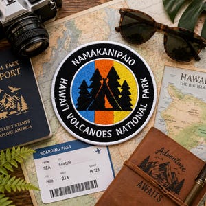May include: A circular embroidered patch featuring a black tent and pine tree silhouette against a vertical blue, orange, and yellow background. The white border text reads NAMAKANIPAIO HAWAI'I VOLCANOES NATIONAL PARK in black lettering.