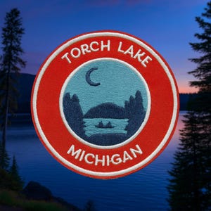 May include: Embroidered patch with a red border and white trim. The patch features the words "TORCH LAKE MICHIGAN" in white, encircling a blue and teal lake scene with a crescent moon and silhouettes of trees and boats.
