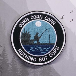 May include: A black and white embroidered patch with a fisherman silhouette casting a line in a blue lake under a full moon. The text around the image reads "Corn Corn Corn Nothing But Corn".