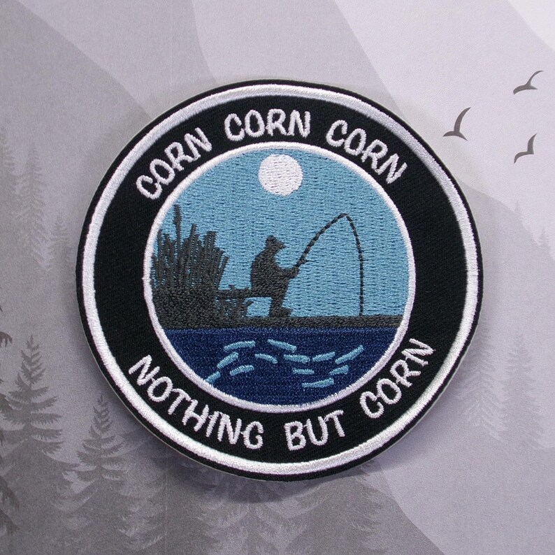 May include: A black and white embroidered patch with a fisherman silhouette casting a line in a blue lake under a full moon. The text around the image reads "Corn Corn Corn Nothing But Corn".