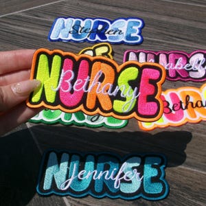 May include: Embroidered patches with the word "NURSE" in various colors and fonts. Some patches include names like "Bethany" and "Jennifer". The patches have a colorful, bubble letter design, with a black border. These patches are likely for medical professionals.