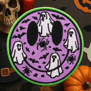 May include: A circular Halloween patch with a purple background and a green border. The patch features a smiley face design with black eyes and mouth, white ghosts, black spiders, and bats.