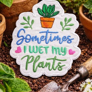 May include: A white embroidered patch with a scalloped edge featuring a potted plant graphic and the text Sometimes I WET MY Plants in blue, green, and pink thread. The design includes small green leaves and two pink hearts on a white fabric base.