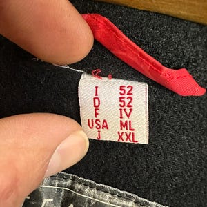 Puede incluir: Primer plano de una etiqueta de ropa con el texto "I 52, D 52, F IV, USA ML, J XXL". La etiqueta es blanca con texto rojo y est&aacute; adherida a una prenda negra. Se ve un lazo de tela roja.