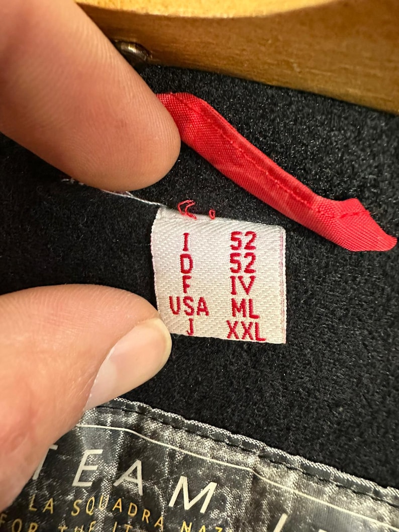 Puede incluir: Primer plano de una etiqueta de ropa con el texto "I 52, D 52, F IV, USA ML, J XXL". La etiqueta es blanca con texto rojo y est&aacute; adherida a una prenda negra. Se ve un lazo de tela roja.