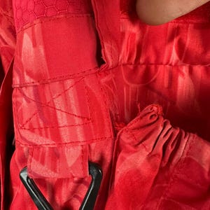 Puede incluir: Primer plano de tela roja brillante con un patr&oacute;n texturizado y herrajes triangulares negros. La tela tiene costuras visibles y una textura de panal en algunas &aacute;reas. La imagen destaca los detalles del material y la construcci&oacute;n.