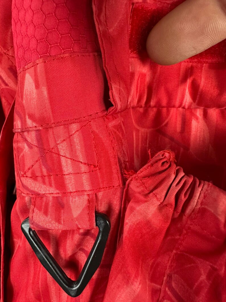 Puede incluir: Primer plano de tela roja brillante con un patr&oacute;n texturizado y herrajes triangulares negros. La tela tiene costuras visibles y una textura de panal en algunas &aacute;reas. La imagen destaca los detalles del material y la construcci&oacute;n.