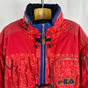 Puede incluir: Chaqueta de esqu&iacute; FILA roja y azul. La chaqueta tiene un cuello alto, una cremallera azul y m&uacute;ltiples bolsillos. El logotipo de FILA es visible en la parte delantera. La chaqueta tiene un patr&oacute;n texturizado.
