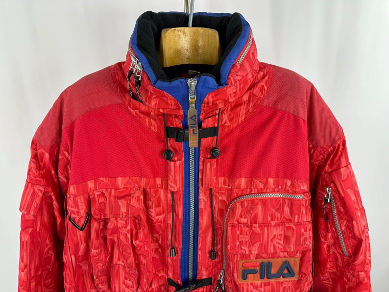 Puede incluir: Chaqueta de esqu&iacute; FILA roja y azul. La chaqueta tiene un cuello alto, una cremallera azul y m&uacute;ltiples bolsillos. El logotipo de FILA es visible en la parte delantera. La chaqueta tiene un patr&oacute;n texturizado.