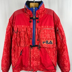 Puede incluir: Chaqueta de esqu&iacute; FILA roja vintage con cuello azul y negro. La chaqueta tiene una cremallera completa, m&uacute;ltiples bolsillos y el logotipo de FILA en el pecho. La chaqueta tiene un dise&ntilde;o estampado y est&aacute; colgada de una percha.