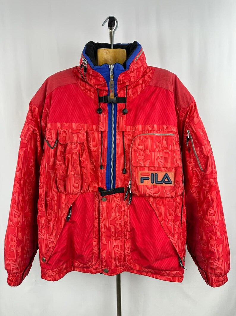 Puede incluir: Chaqueta de esqu&iacute; FILA roja vintage con cuello azul y negro. La chaqueta tiene una cremallera completa, m&uacute;ltiples bolsillos y el logotipo de FILA en el pecho. La chaqueta tiene un dise&ntilde;o estampado y est&aacute; colgada de una percha.