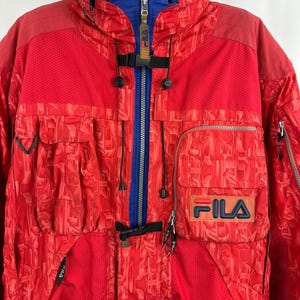 Puede incluir: Chaqueta de esqu&iacute; FILA roja y azul con un patr&oacute;n geom&eacute;trico. La chaqueta presenta una cremallera azul, un cuello azul y el logotipo de FILA en el pecho. Tiene m&uacute;ltiples bolsillos y un cuello alto.
