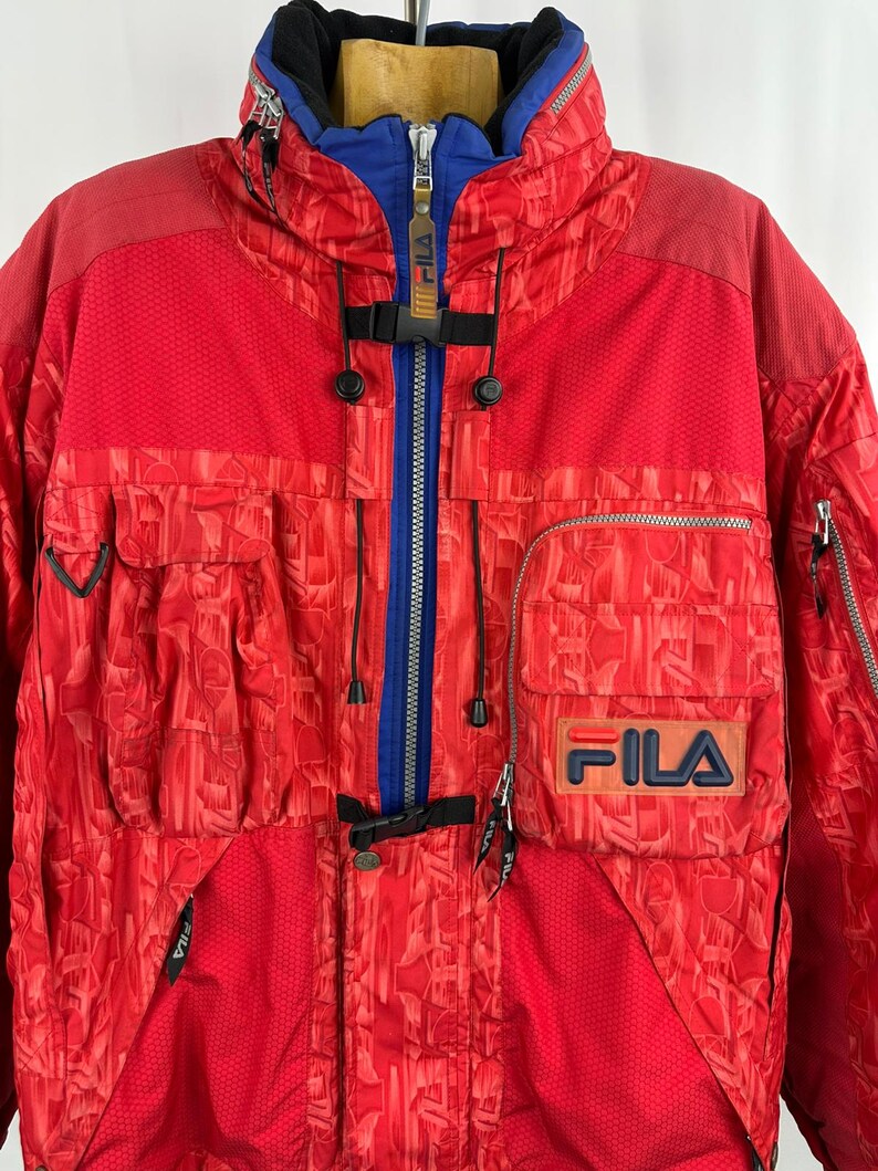 Puede incluir: Chaqueta de esqu&iacute; FILA roja y azul con un patr&oacute;n geom&eacute;trico. La chaqueta presenta una cremallera azul, un cuello azul y el logotipo de FILA en el pecho. Tiene m&uacute;ltiples bolsillos y un cuello alto.