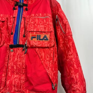 Puede incluir: Chaqueta de esqu&iacute; Fila roja con un patr&oacute;n geom&eacute;trico. La chaqueta presenta una cremallera, cuello y detalles en azul, junto con m&uacute;ltiples bolsillos y el logotipo de Fila en el pecho. La chaqueta tiene un cuello alto y un cierre de hebilla negro.