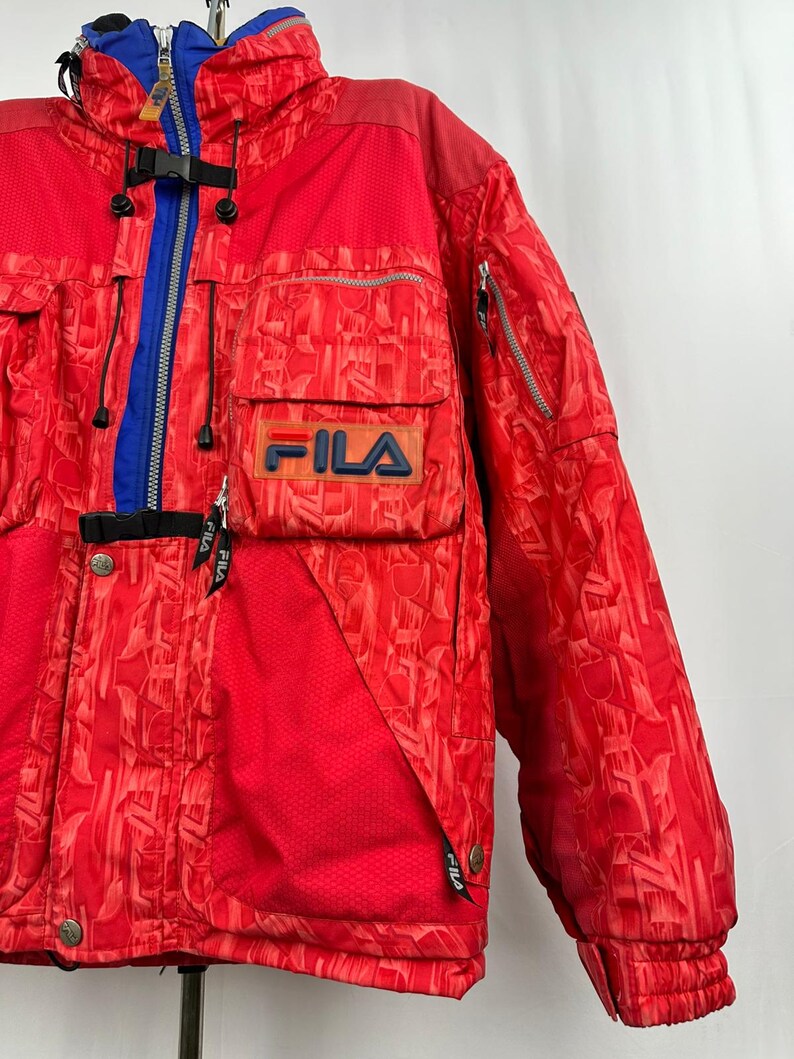 Puede incluir: Chaqueta de esqu&iacute; Fila roja con un patr&oacute;n geom&eacute;trico. La chaqueta presenta una cremallera, cuello y detalles en azul, junto con m&uacute;ltiples bolsillos y el logotipo de Fila en el pecho. La chaqueta tiene un cuello alto y un cierre de hebilla negro.