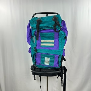 Può includere: Uno zaino da trekking vintage verde acqua, viola e nero con telaio in metallo. Lo zaino presenta più scomparti, cinghie e una piccola toppa con una scena naturalistica. Il design include strisce orizzontali e una base nera.