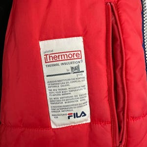 Puede incluir: Primer plano del interior de una chaqueta roja con una etiqueta blanca. La etiqueta dice "Thermore Thermal Insulation" y contiene texto en varios idiomas. Una cremallera roja recorre verticalmente. Fabricado para Fila.