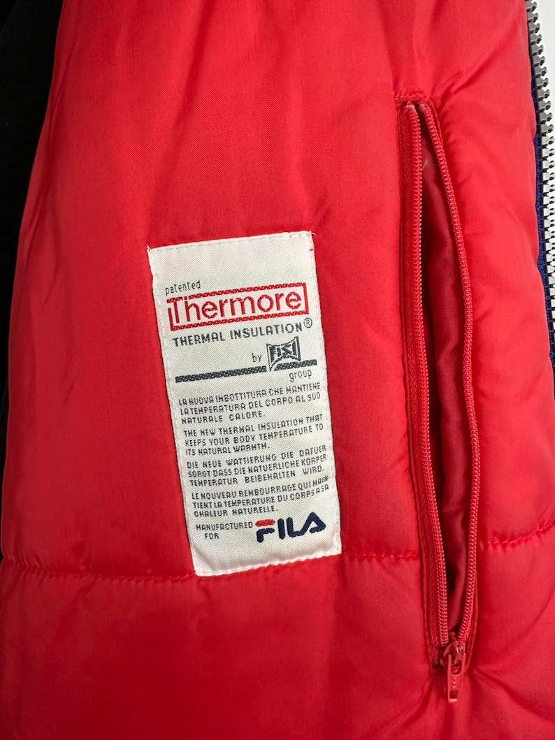 Puede incluir: Primer plano del interior de una chaqueta roja con una etiqueta blanca. La etiqueta dice "Thermore Thermal Insulation" y contiene texto en varios idiomas. Una cremallera roja recorre verticalmente. Fabricado para Fila.